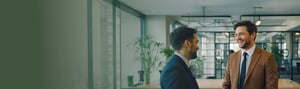 Zwei Männer im Anzug lachen in einem modernen Büro, symbolisieren erfolgreiche Anlageberatung und Vermögensverwaltung.